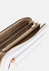 Portafoglio bianco con hardware dorato, chiusura a zip e più scomparti. L'interno presenta una fodera liscia beige e una sezione decorata.