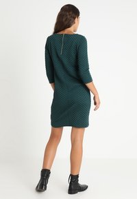 Vestido acolchado en verde azulado con mangas tres cuartos, que presenta una cremallera en la parte posterior y una silueta ajustada. Combinado con botas negras hasta el tobillo.