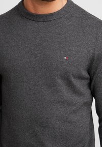 Pull en maille grise avec un col rond et des poignets côtelés, présentant un petit logo rouge, blanc et bleu sur le côté gauche de la poitrine.