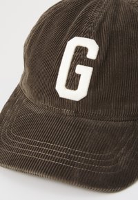 Brown corduroy baseball cap with a white fabric "G" logo on the front, featuring stitched detailing and a curved visor.