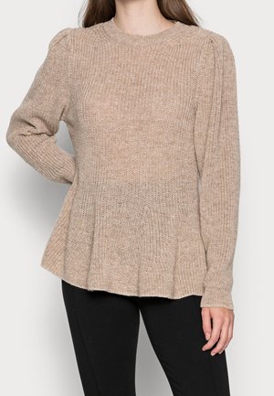 Femme portant un pull beige en maille à manches longues avec des épaules légèrement bouffantes et un pantalon noir, debout devant un fond uni.