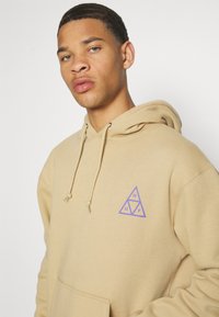Sudadera beige hecha de tela suave, con un logo triangular morado en el lado izquierdo del pecho, con cordones y un bolsillo frontal.