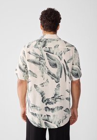 Camisa de manga corta en tejido ligero, con fondo color crema y estampado de hojas tropicales verdes, y dobladillo relajado y curvado.