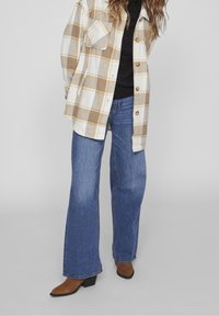 Bílé flanelové košile s hnědou kostičkou, tmavý pletený top pod ní, široké modré džíny a hnědé boty na podpatku. Ležérní vrstvený outfit.