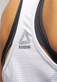 Soutien-gorge de sport blanc et noir avec une texture en maille, présentant un logo Reebok gris et un design triangulaire au dos.