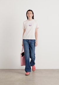 Maglietta bianca in cotone con un logo viola, jeans ampi blu e scarpe da ginnastica rosa. Il modello porta una borsa a spalla rosa con un interno nero.