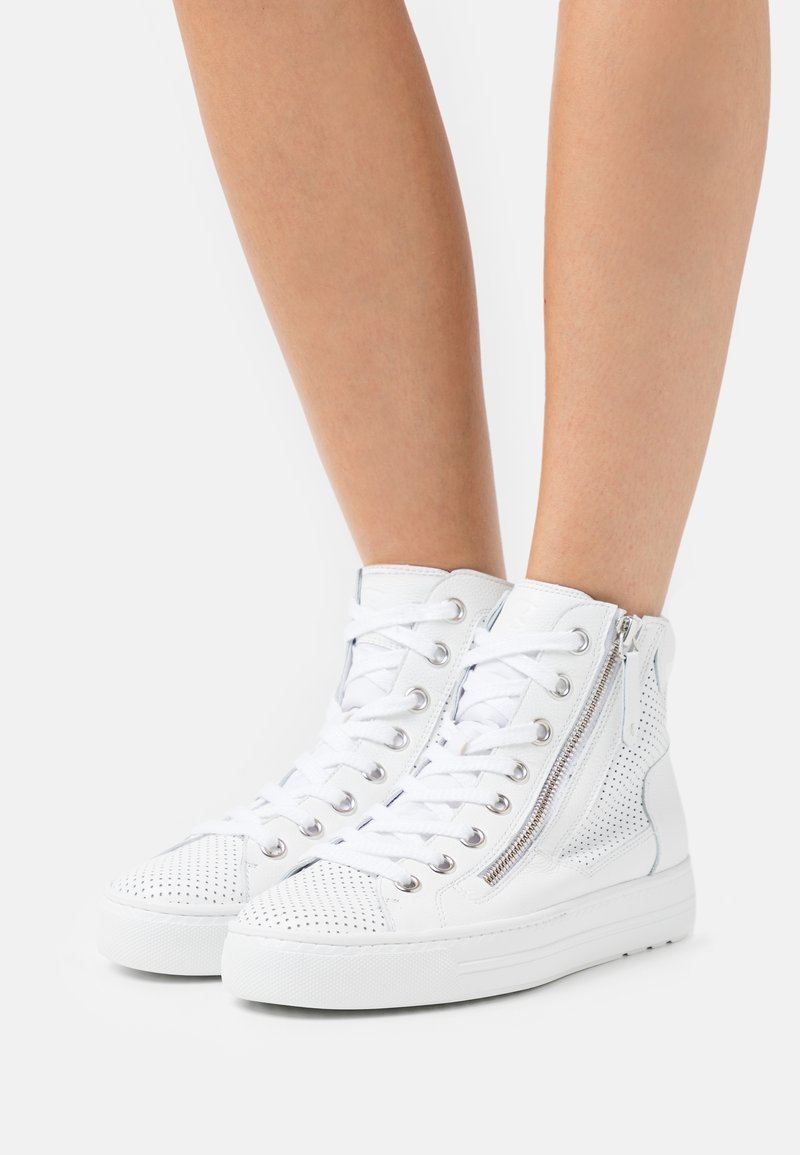 Legs wearing white high-top sneakers with perforated toe and side panels, white laces, and silver side zippers on a white background.