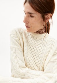 Gebreide crèmekleurige trui met kabel- en textuurpatronen, met een hoge kraag en een ontspannen pasvorm, gelaagd over een gestructureerde halsdoek.