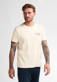 Krémové tričko s krátkým rukávem a kulatým výstřihem, s malým potiskem loga na levém prsu. Hladká bavlněná látka, volný střih.