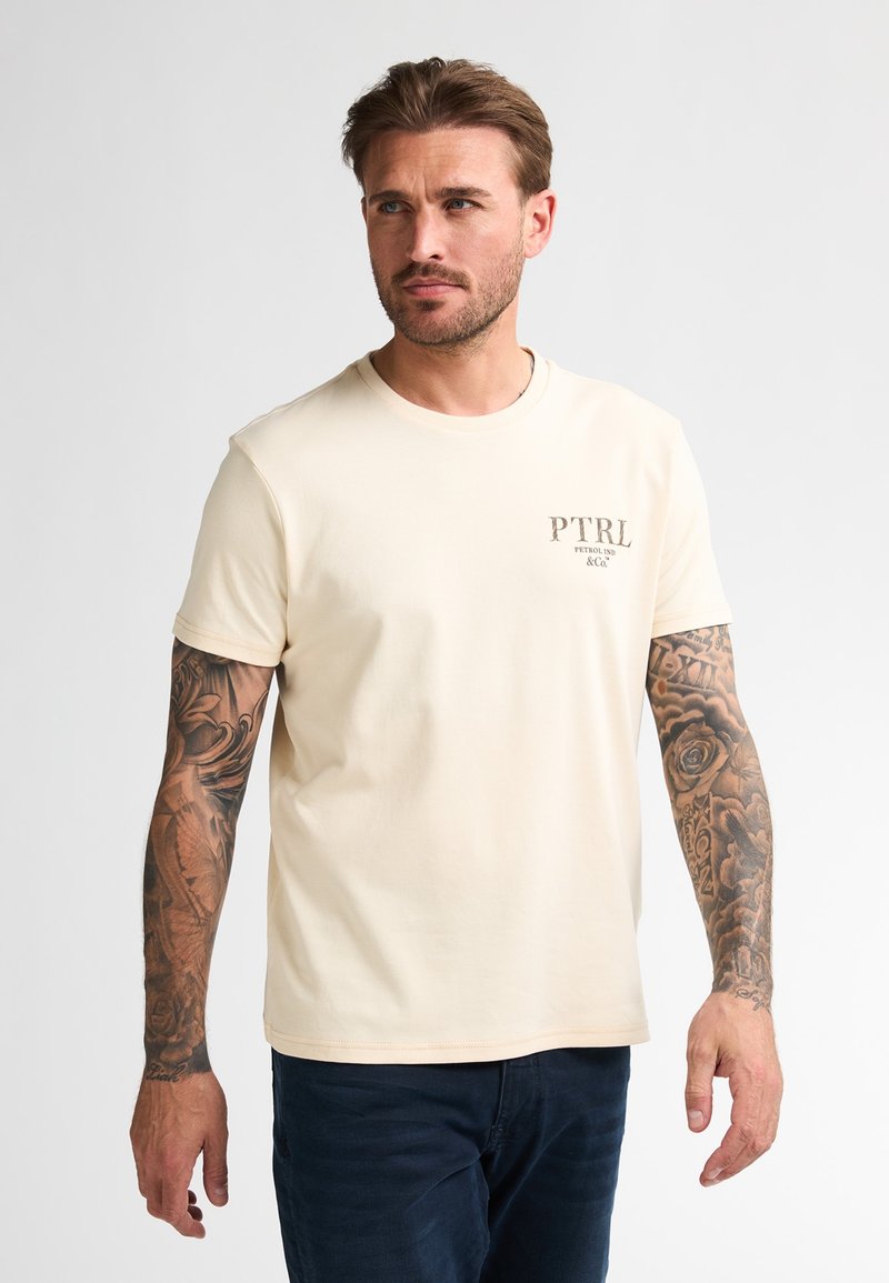 Krémové tričko s krátkým rukávem a kulatým výstřihem, s malým potiskem loga na levém prsu. Hladká bavlněná látka, volný střih.