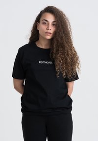 Sort bomulds t-shirt med et hvidt "PENTHOUSE" logo på brystet, med korte ærmer og en afslappet pasform. Sammen med sorte bukser.