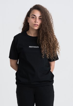 Sort bomulds t-shirt med et hvidt "PENTHOUSE" logo på brystet, med korte ærmer og en afslappet pasform. Sammen med sorte bukser.