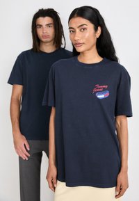 Camiseta de algodón azul marino con cuello redondo y mangas cortas, que presenta un gráfico en rojo y azul de "Tommy Jeans". Acabado de tela texturizada.