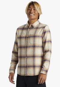 Camisa de cuadros de manga larga en crema con acentos en púrpura y marrón, que presenta dos bolsillos frontales y cierre de botones. Fabricada en un tejido suave y texturizado.