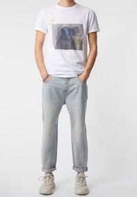 Bílý bavlněný tričko s grafickým potiskem, spárované s světlými džíny. Boty jsou béžové s texturovanou podrážkou. Volný střih.