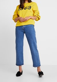 Felpa di cotone gialla con la scritta "1969" stampata in nero, abbinata a jeans in denim azzurro a gamba dritta e scarpe slip-on nere.