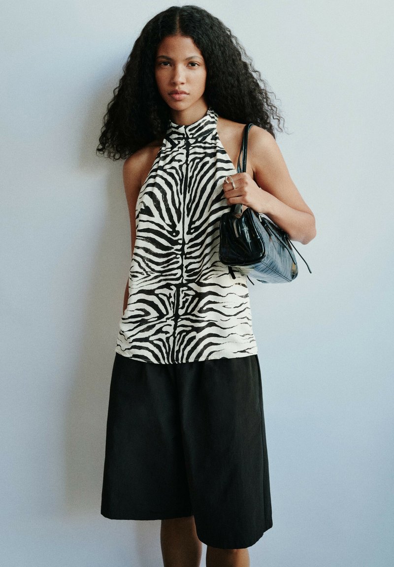 Jeune femme aux cheveux bouclés portant un haut dos nu à imprimé zèbre et un short noir à jambes larges, tenant un petit sac à main noir sur l'épaule.