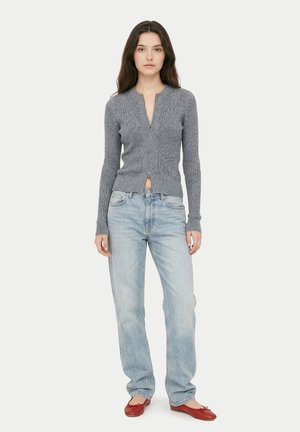 Frau steht und trägt einen grauen, gerippten Pullover mit Reißverschluss, hellblaue hoch taillierte Jeans und rote Ballerinas vor einfachem Hintergrund.