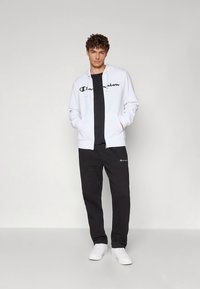 Noor mees seisab käed taskutes, kandes valget lukuga pusa, musta t-särki, musti dressipükse ja valgeid tossusid tavalise tausta ees.