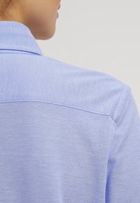 Hellblaues, strukturiertes Hemd mit Kragen, das ein nahtloses Rückendesign und sichtbare Nähte entlang der Schultern und Halslinie aufweist.