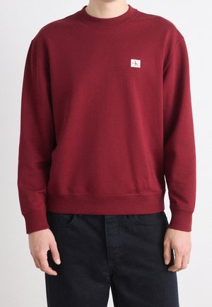 Osoba ubrana w jednolity bordowy długi rękaw bluzy z małą białą naszywką z logo Calvin Klein na lewej piersi, zestawioną z czarnymi spodniami.