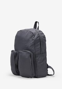 Mochila de nailon gris con dos bolsillos frontales con cremallera, espalda acolchada y correas ajustables. Textura suave y ligera con forma redondeada.