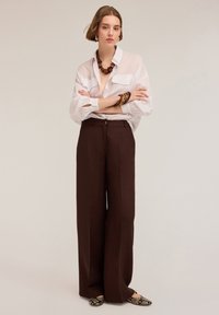Camasa albă, cu nasturi, dotată cu două buzunare pe piept, asortată cu pantaloni maro cu talie înaltă și lărgime. Accesoriile includ un colier masiv și brățări.