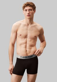 Boxers en coton noir avec une ceinture élastique grise portant le logo "Calvin Klein". Le modèle a une silhouette mince, sur fond gris.