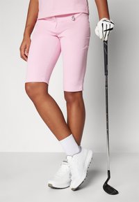 Rózsaszín golfnadrág sima textúrával és felgombolt szegéssel, fehér sportcipővel és golfkesztyűvel párosítva, egy klub mellett állva.