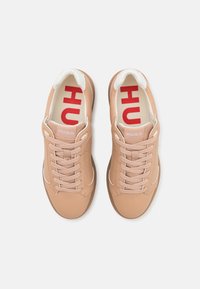 Zapatillas beige con parte superior de cuero liso, acentos en blanco y suela con textura; cuentan con cordones planos y una lengüeta con logo rojo.