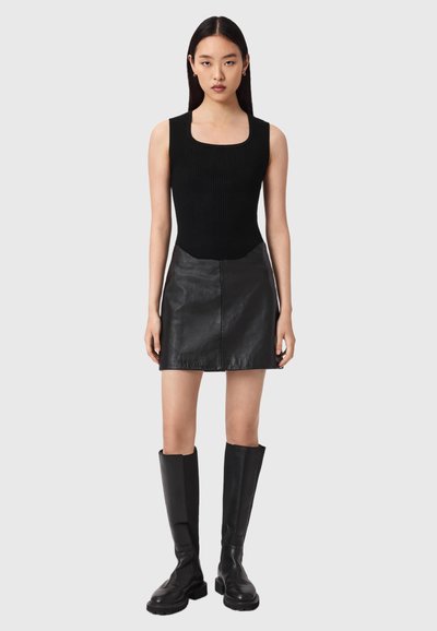 Jeune femme debout devant un fond uni, portant un haut noir sans manches, une mini-jupe en cuir noir et des bottes noires montantes jusqu'aux genoux.
