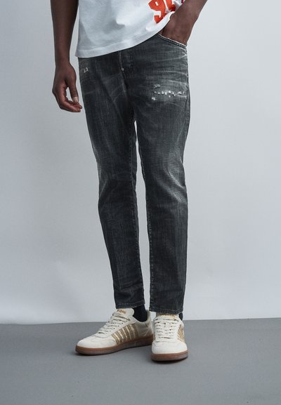 Personne portant un jean slim noir délavé et des baskets blanches avec des bandes dorées, debout avec une main dans la poche devant un fond uni.