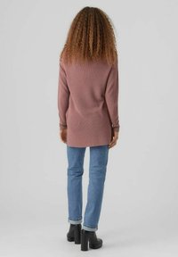 Personne aux cheveux bouclés portant un long pull mauve, un jean bleu retroussé et des bottines à plateforme noires, debout devant un fond gris uni.
