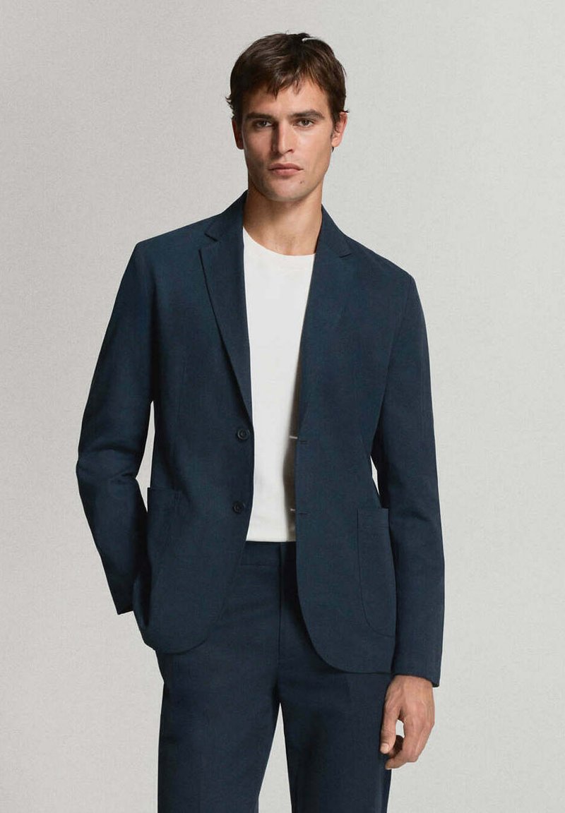 Homme portant une veste de costume bleu foncé et un pantalon assorti avec une chemise blanche unie, debout devant un fond neutre et clair.