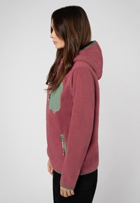 Fleece hoodie in donkerroze met een groene accentzak. Heeft een capuchon, ritssluitingen en een zachte, textuur oppervlak.