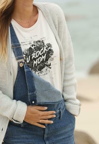 T-shirt graphique blanc avec le texte "U ROCK MOM" sous un cardigan tricoté gris clair. Des salopettes en denim, bleu avec des accessoires dorés, affichent une coupe décontractée.