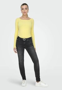 Geel geribbelde top met lange mouwen en een vierkante halslijn, gecombineerd met zwarte skinny jeans en witte sneakers. De riem heeft een cirkelvormige zilveren hardwareaccent.