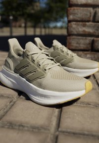 Beige atletiske sneakers med en struktureret mesh-overdel, tre grønne striber, en tyk hvid mellemsål og en gul gummisål.