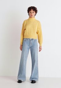 Pulover cu guler înalt tricotat în stil cable-knit de culoare muștar, asortat cu blugi de tip wide-leg de culoare albastră deschisă și pantofi negri. Textură fină și croială lejeră.