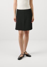 Jupe grise foncée, longueur genoux, avec une texture lisse et une taille ajustée. Portée avec des ballerines en cuir verni noires.