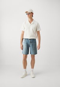 Gaišs, ar tekstūru apstrādāts balts īsām piedurknēm krekls, zili džinsu šorti, baltas kedas un bēša cepure, ar taisnstūra saulesbrillēm.