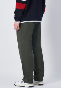 Khakigrüne weite Hose aus Baumwolle mit Seitennähten und lässigem Schnitt, kombiniert mit weißen Turnschuhen. Keine auffälligen Muster oder Akzente.