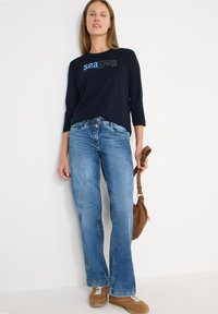 Chemise bleu marine à manches longues avec motif "mer", jean bleu à jambes évasées, sac marron et baskets beige. Tissu lisse et coupe décontractée.