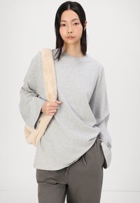 Chemise oversize grise à manches longues avec des manches larges, associée à un pantalon gris ample, tenant une sangle en fausse fourrure crème.