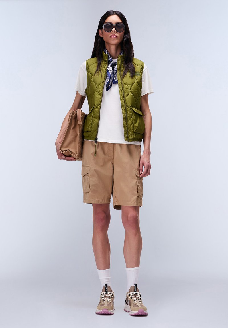 Oliwkowa, pikowana kamizelka, biała koszulka, beżowe spodenki cargo, brązowa torba, białe skarpetki, beżowo-różowe sneakersy. Eleganckie okulary przeciwsłoneczne, związany szalik.