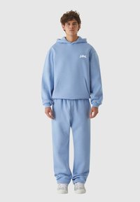 Šviesiai mėlynas džemperis derinamas su atitinkančiomis kelnėmis. Džemperis turi priekinę kišenę ir gobtuvą; glotni, švelni tekstūra. Baltas logotipas kaip akcentas.