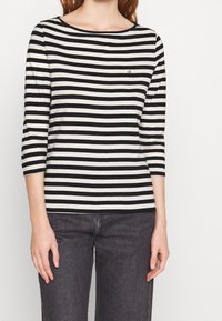 Svartvit randig långärmad skjorta med rund halsringning och liten "ck"-logotyp på bröstet, tillsammans med mörkgrå jeans.