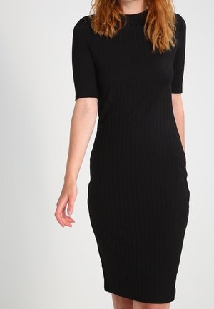 Kobieta o rudych włosach ubrana w dopasowaną czarną prążkowaną sukienkę midi z krótkimi rękawami, stojąca na jednolitym jasnym tle.