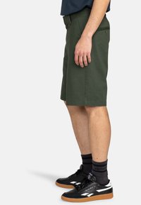 Dunkelgrüne Shorts mit geradem Schnitt, zwei seitlichen Taschen und einer Gesäßtasche. Kombiniert mit schwarzen Turnschuhen und dunkelgestreiften Socken.