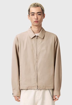 Beige jakke med krave, lynlås og lange ærmer. Stoffet virker glat med minimal syning og sidelommer.